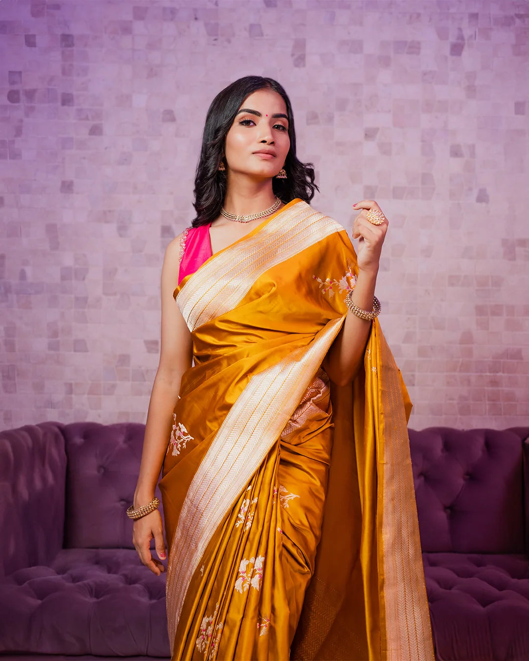 A woman in a gold saree stands confidently in front of a textured wall. She wears jewelry and light makeup