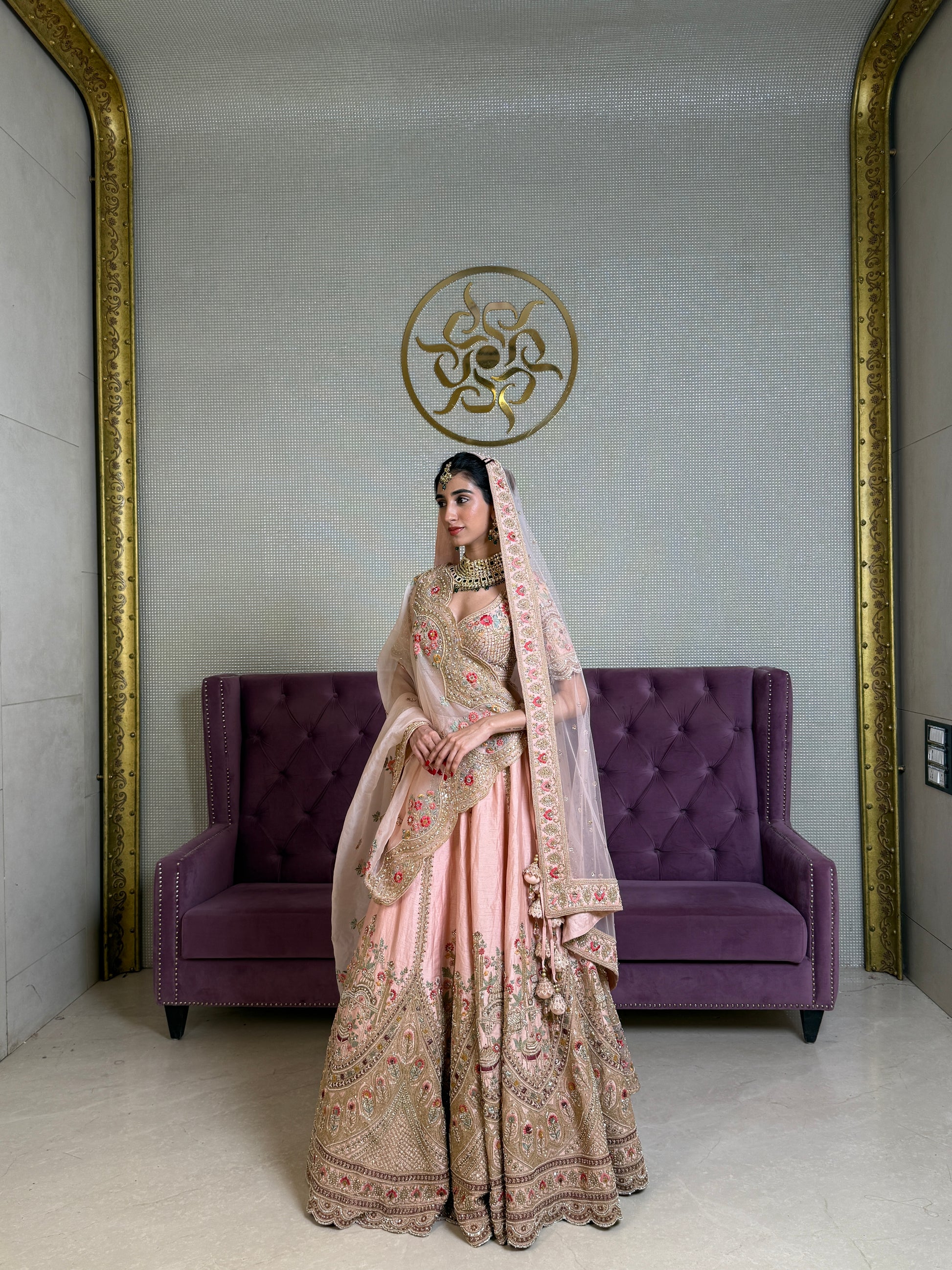A woman in an elegant traditional outfit stands against a wall. She wears an ornate bridal lehenga and dupatta