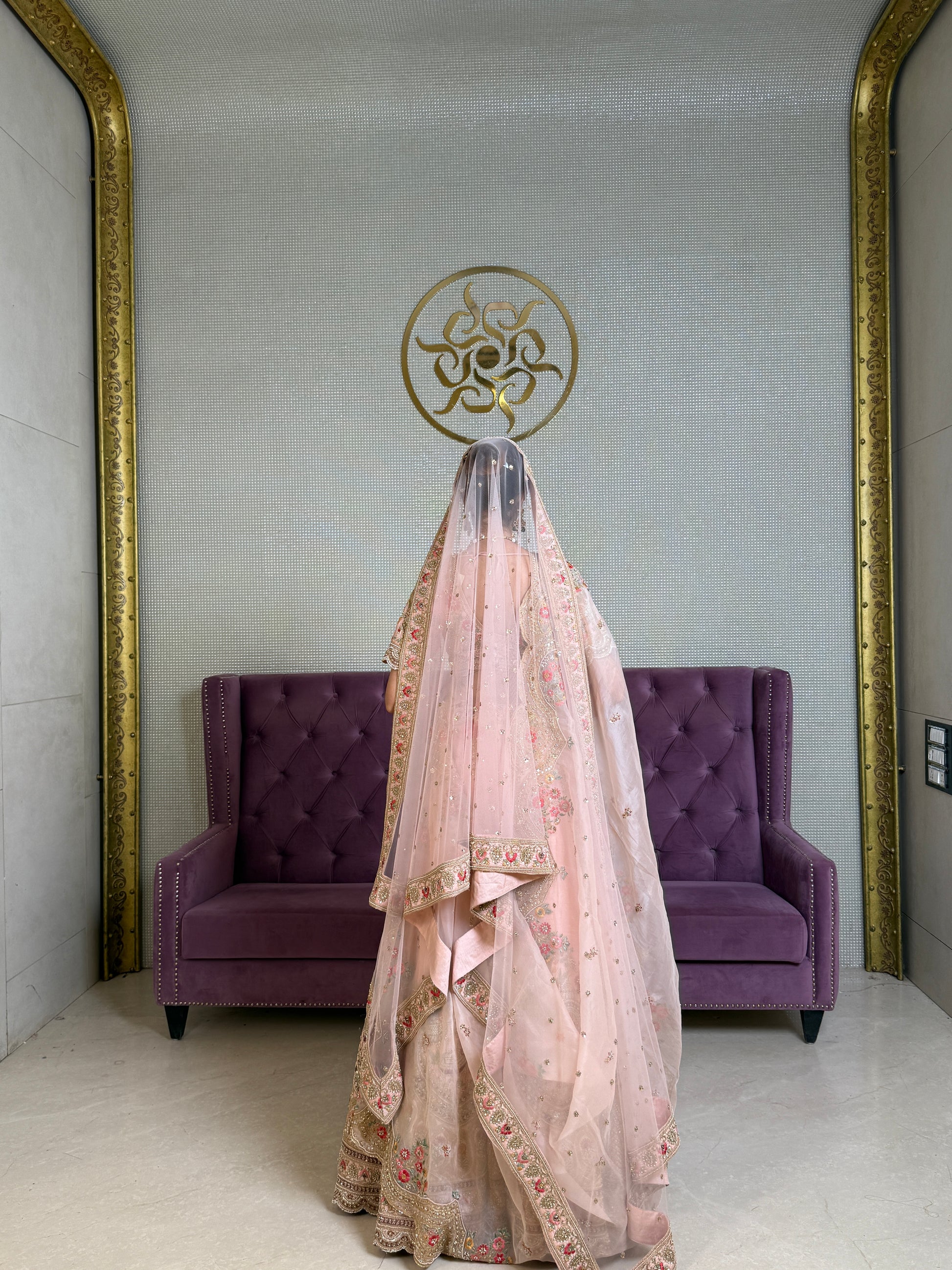 Bride in ornate, pink embroidered silk bridal lehenga stands before a purple tufted sofa