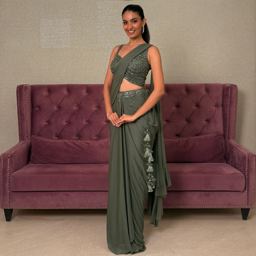 Woman holding a green saree in front of a purple couch with a decorative wall design.
