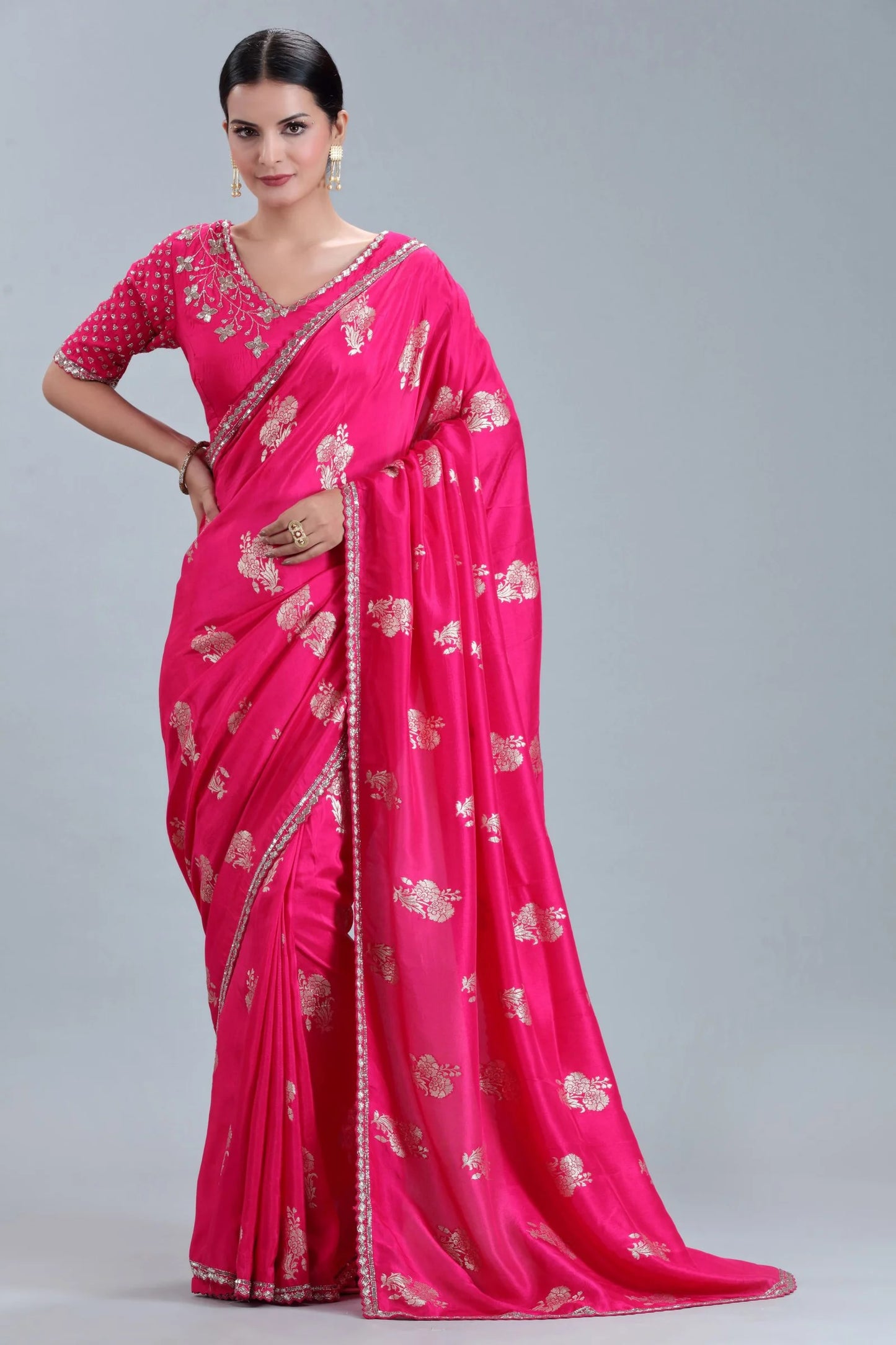 A woman in a vibrant pink saree with intricate gold embroidery and a matching blouse poses against a plain gray background.
