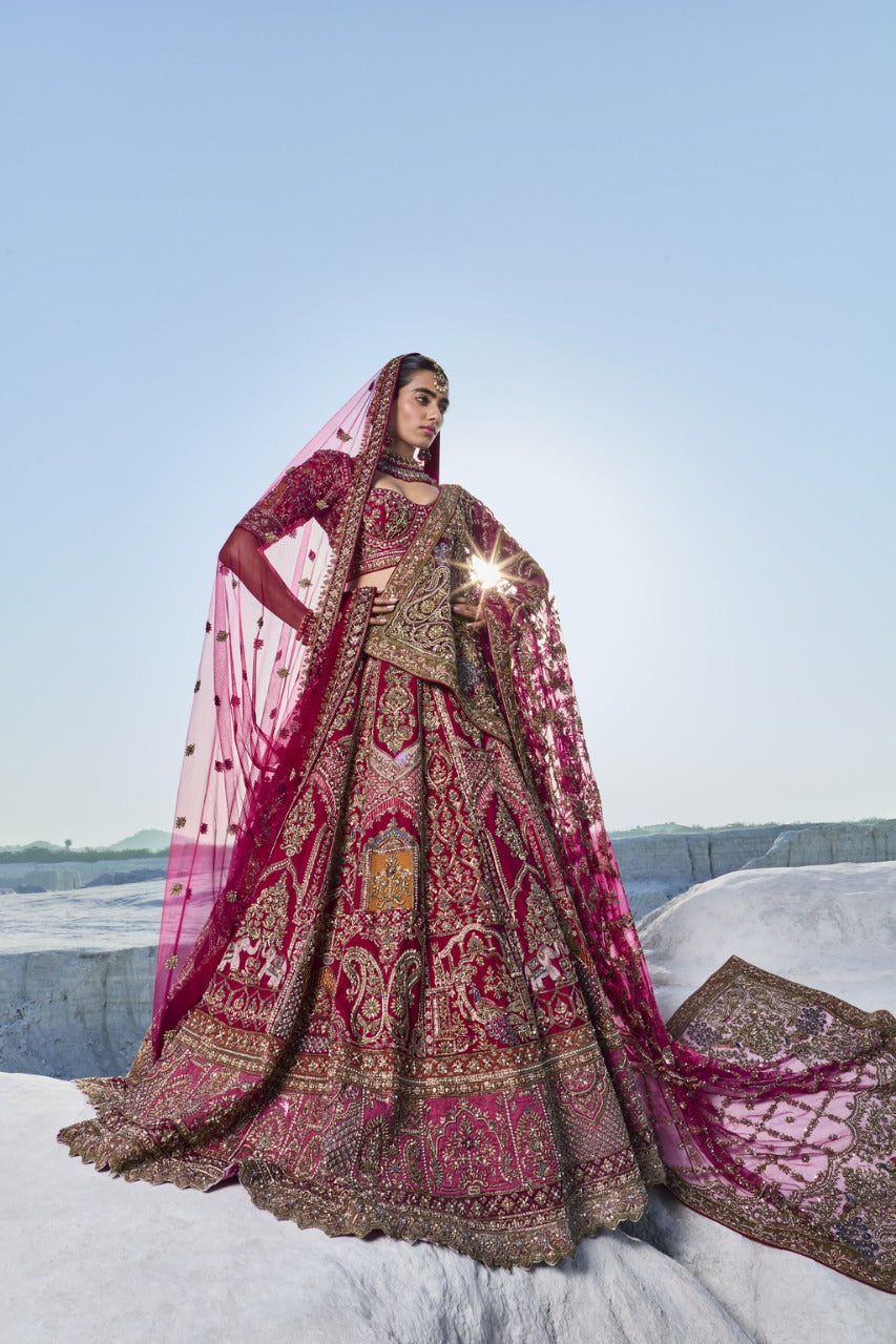 A woman stands against a clear sky, wearing a luxurious, deep pink lehenga with intricate gold embroidery