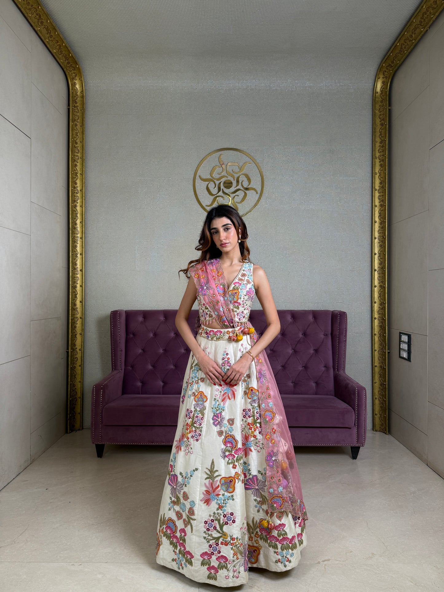 Woman in a Beige floral lehenga set standing in a room with a purple couch and decorative wall.