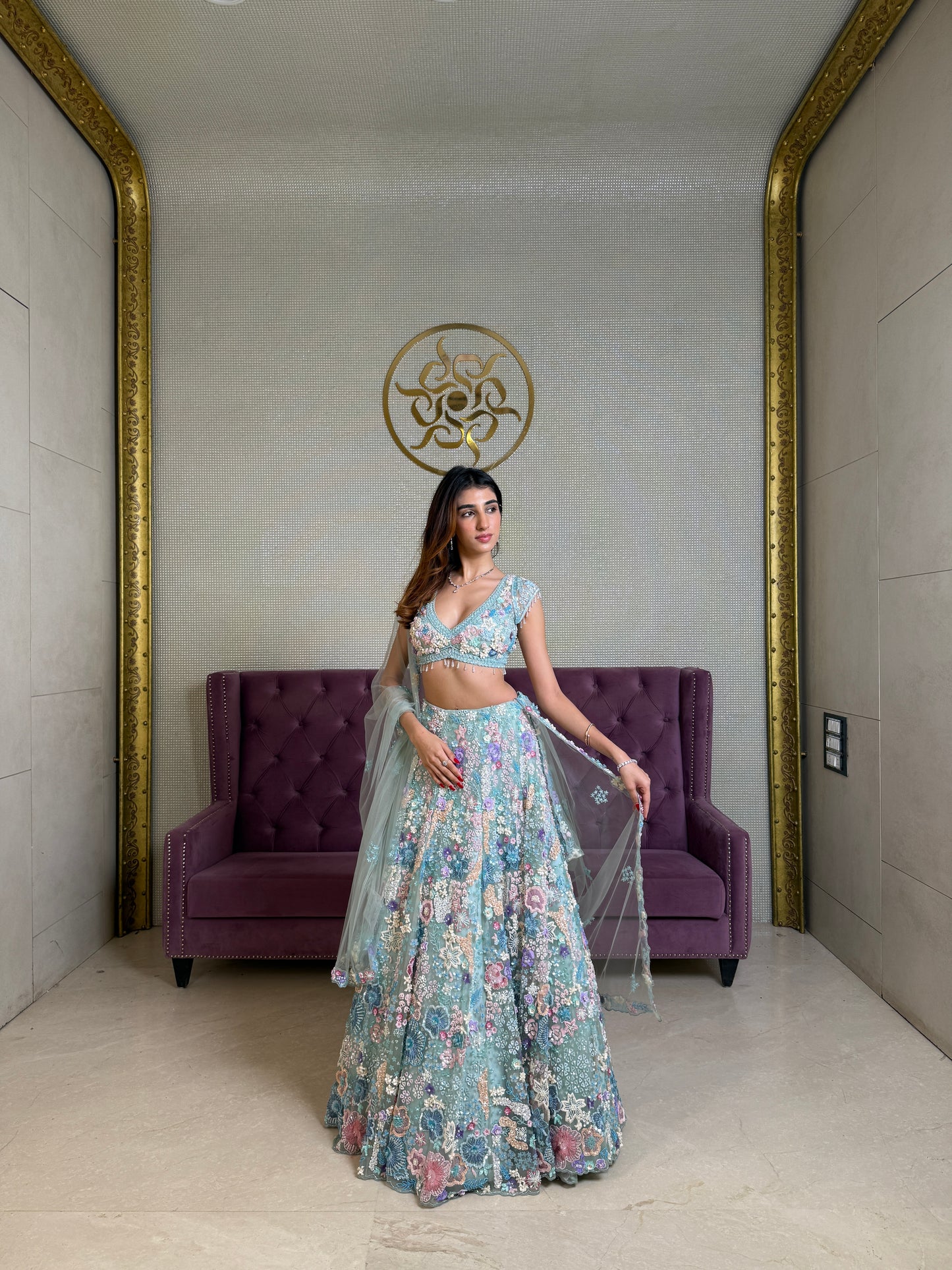 Woman wearing a floral blue lehenga in a room with a purple couch and decorative wall.