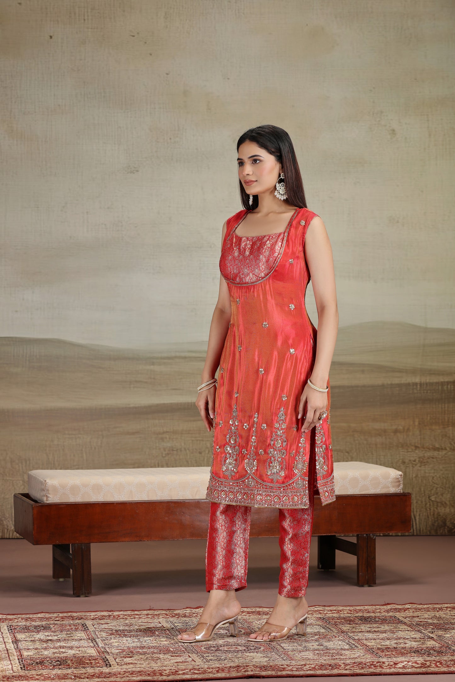 Woman wearing a red traditional outfit with intricate patterns in a room with a textured wall and wooden bench.