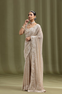 Woman wearing a beige saree with intricate patterns against a green background