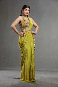 A woman in a vibrant olive saree with colorful embroidery on the blouse poses confidently.