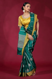 A woman in a vibrant green and gold sari poses elegantly against a deep red backdrop. She wears a golden blouse and large, dangling earrings