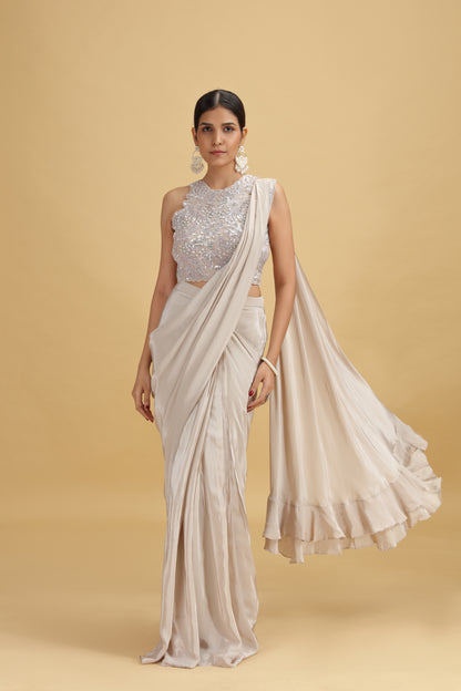 A woman stands against a beige background, wearing a shimmering silver top and a flowing cream saree. She exudes elegance, with her hair tied back and large, ornate earrings.