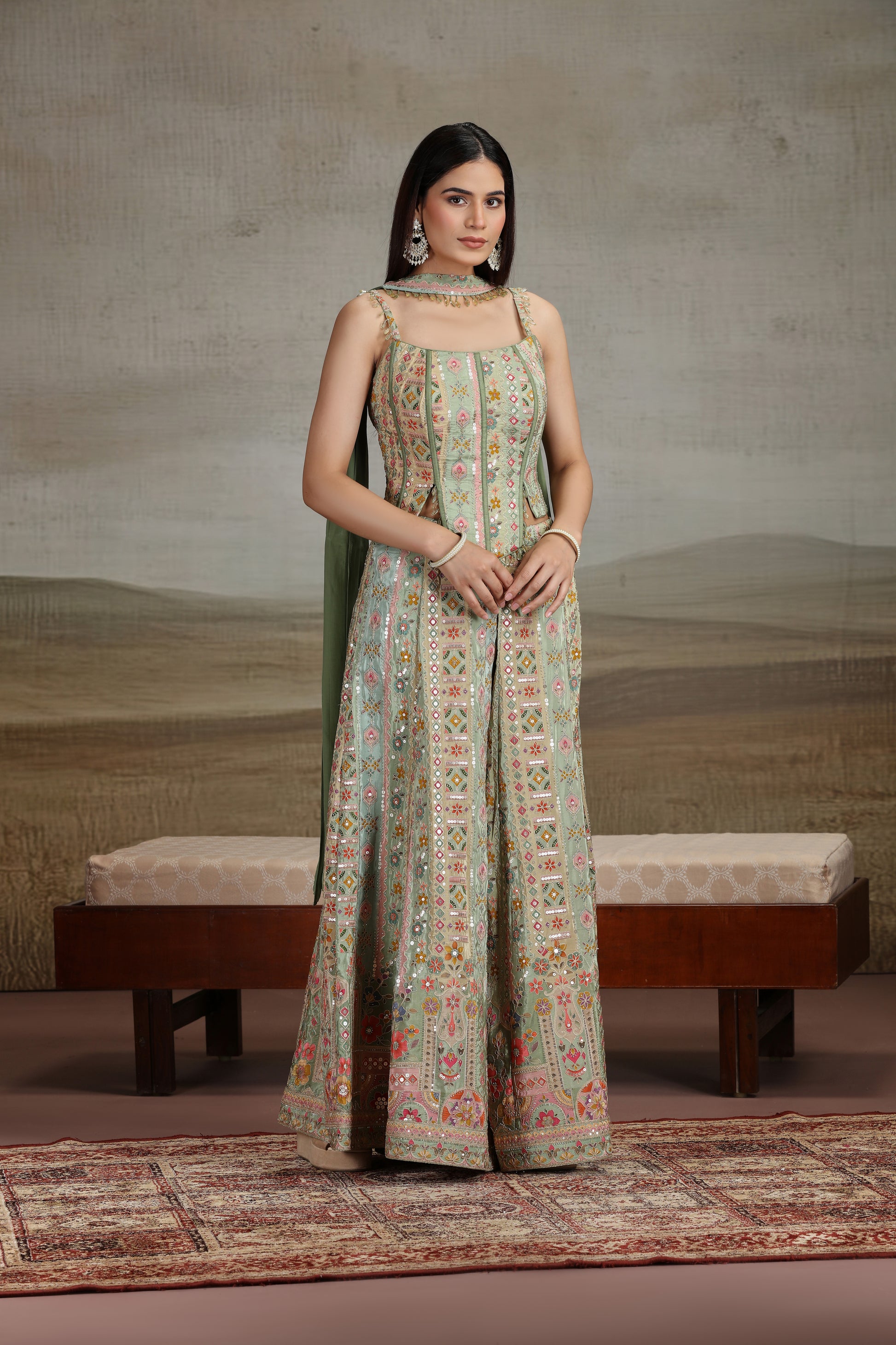 Woman wearing a green floral salwar suit standing in a room with a textured wall and bench.