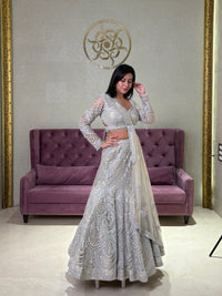 Woman in a silver grey lehenga set standing in a room with a purple couch and decorative wall.