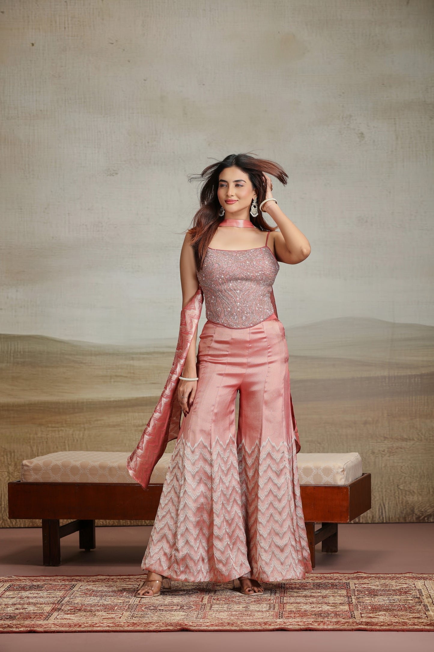 Woman in a peach banarasi suit standing in a room with a textured wall and wooden bench.