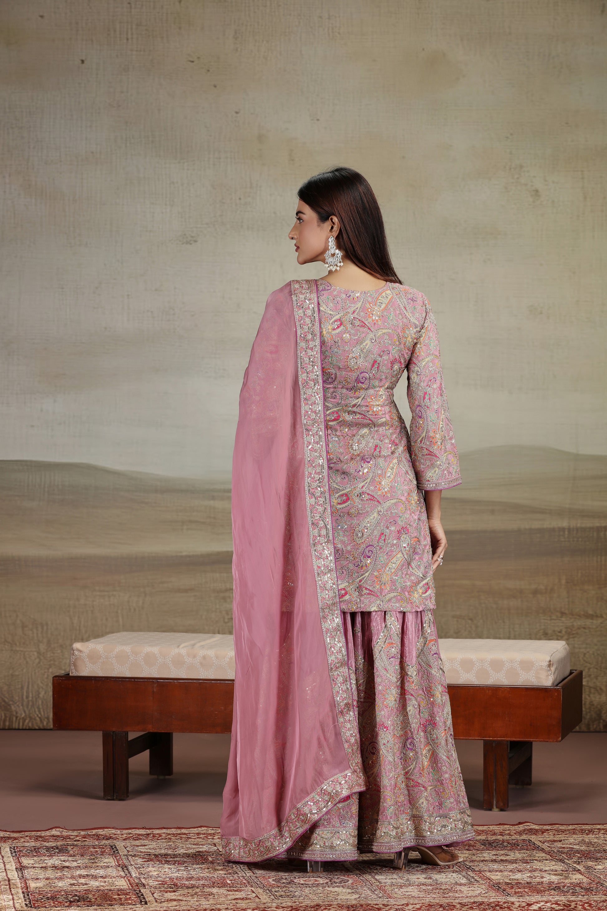Woman in a pink traditional outfit standing in a room with a textured wall and wooden bench.