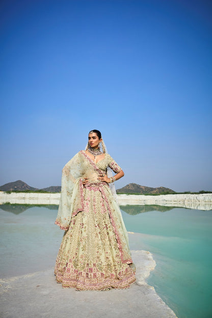 Woman in traditional green raw lehenga standing by a lake with mountains in the background