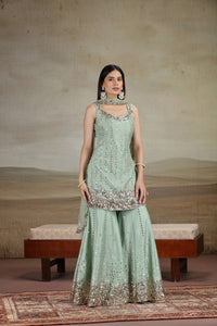 Woman wearing a light green traditional outfit with intricate patterns on a textured wall background