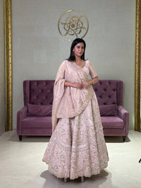 Woman in a blush pink lehenga standing in a room with a purple couch and decorative wall.