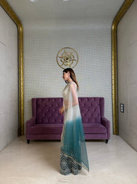 Woman in a long, light-colored dress standing in a room with a purple couch and decorative wall.