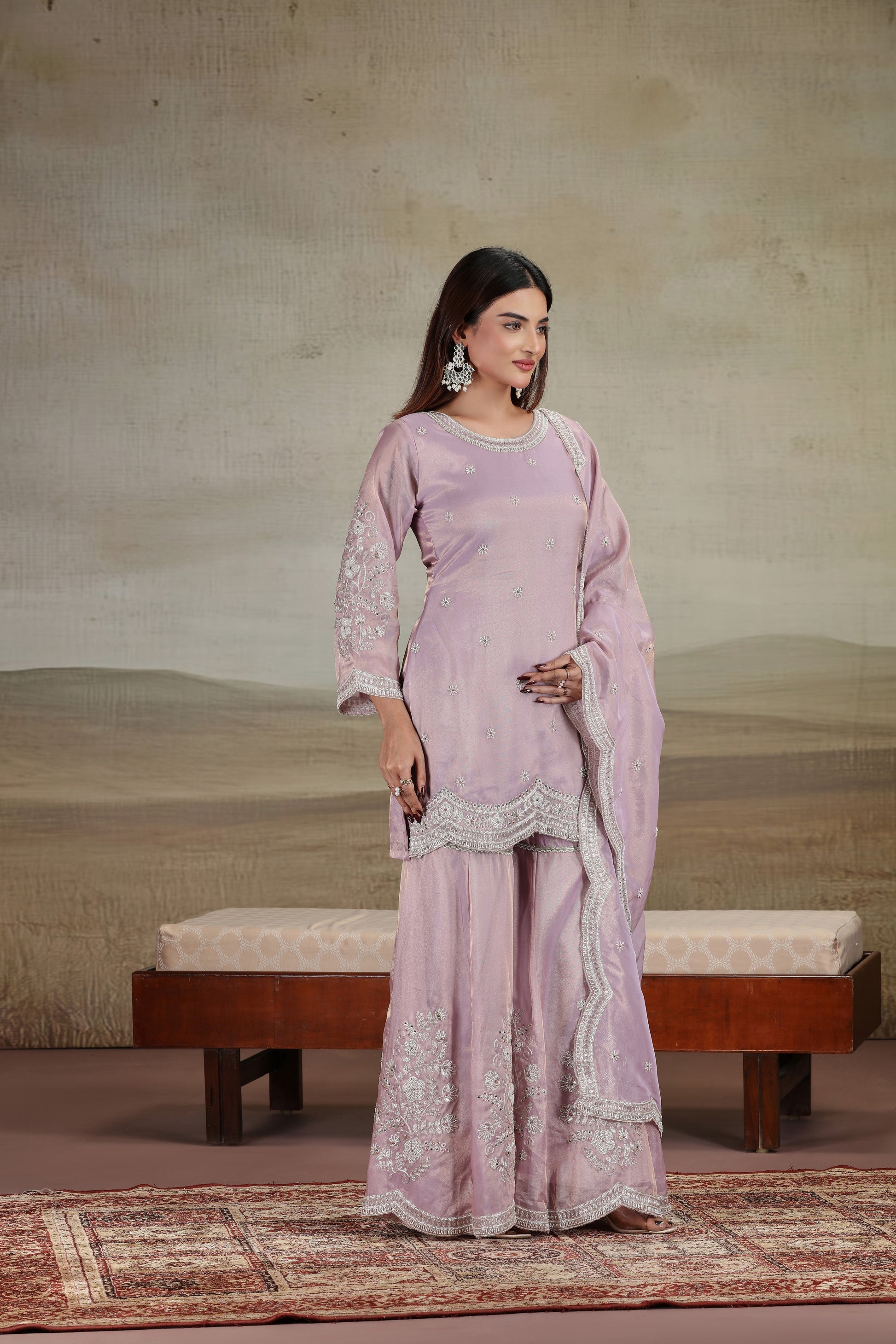 Woman in a lilac traditional outfit with lace details standing in a room with a textured wall and wooden bench.