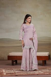 Woman in a lilac traditional outfit with lace details standing in a room with a textured wall and wooden bench.