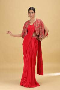 Woman wearing a red saree with a matching blouse and floral jacket on a beige background