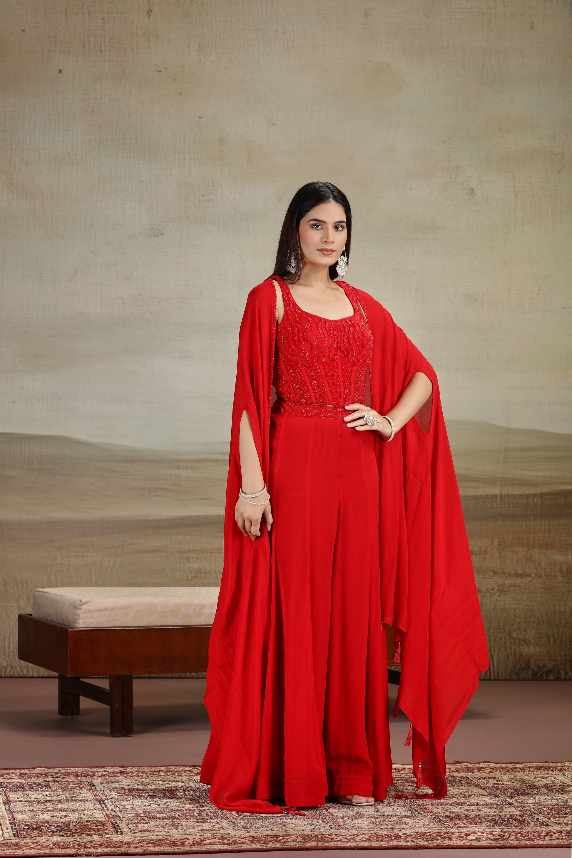 Woman in a designer red salwar suit with intricate cutdana and pearl work standing against a beige wall.