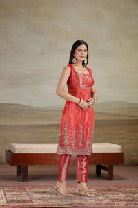 Woman in a rust orange banarsi suit standing in front of a textured wall with a bench.