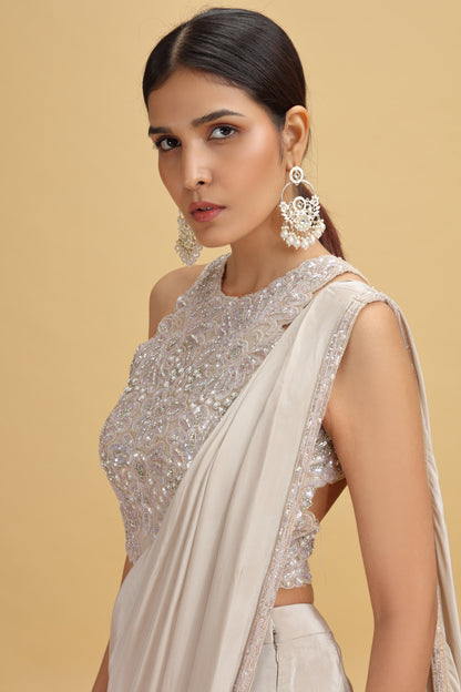Woman in an elegant cream sari with intricate beadwork, large ornate earrings, and sleek hairstyle, set against a soft beige background.