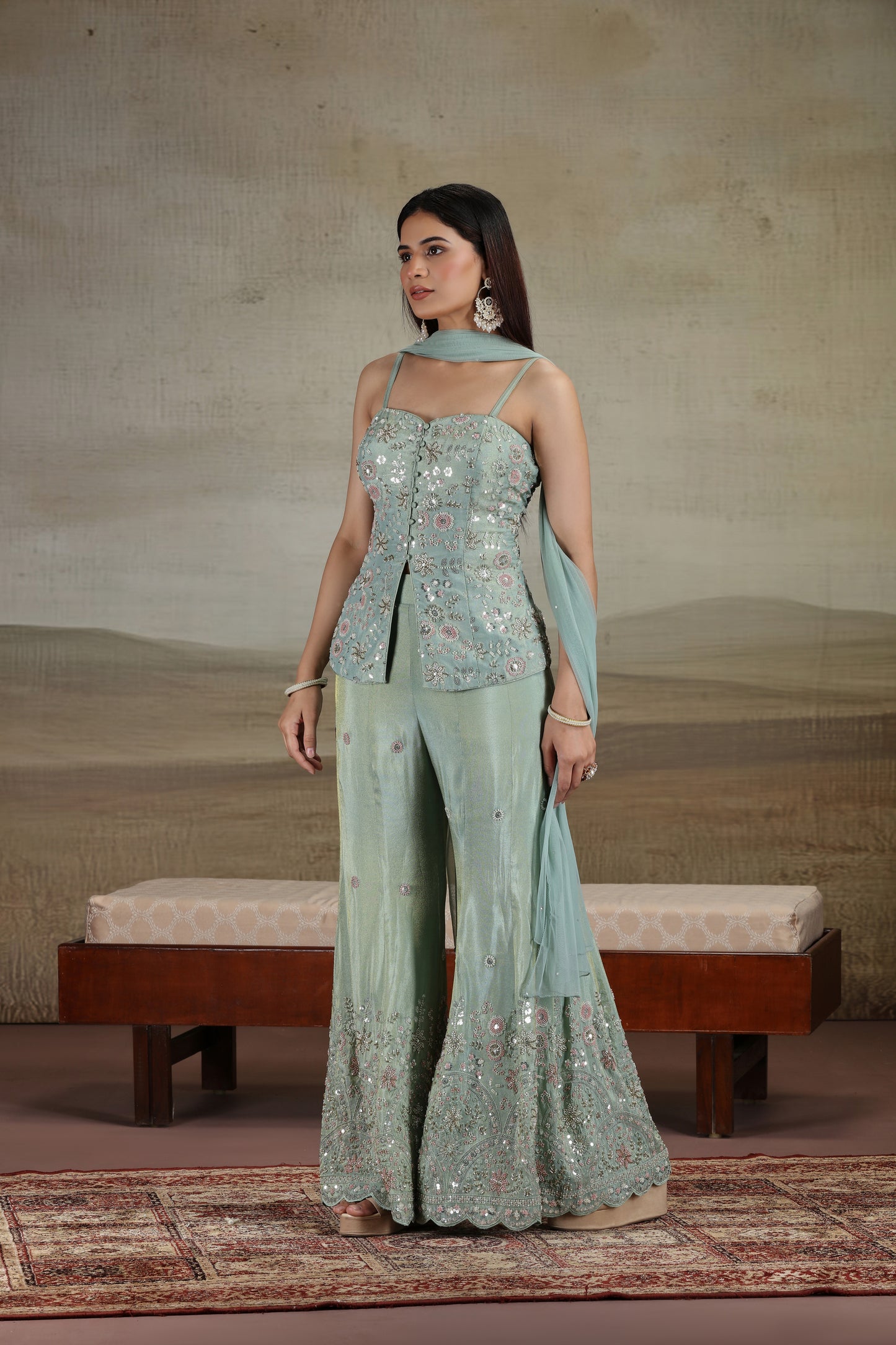 Woman in a pista green tissue salwar suit with sequins thread and cutdana work standing in a room with a textured wall and wooden bench.