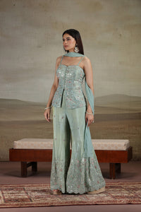 Woman in a pista green tissue salwar suit with sequins thread and cutdana work standing in a room with a textured wall and wooden bench.