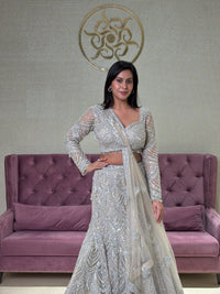 Woman in a grey net lehenga set standing next to a purple couch with a decorative wall in the background.