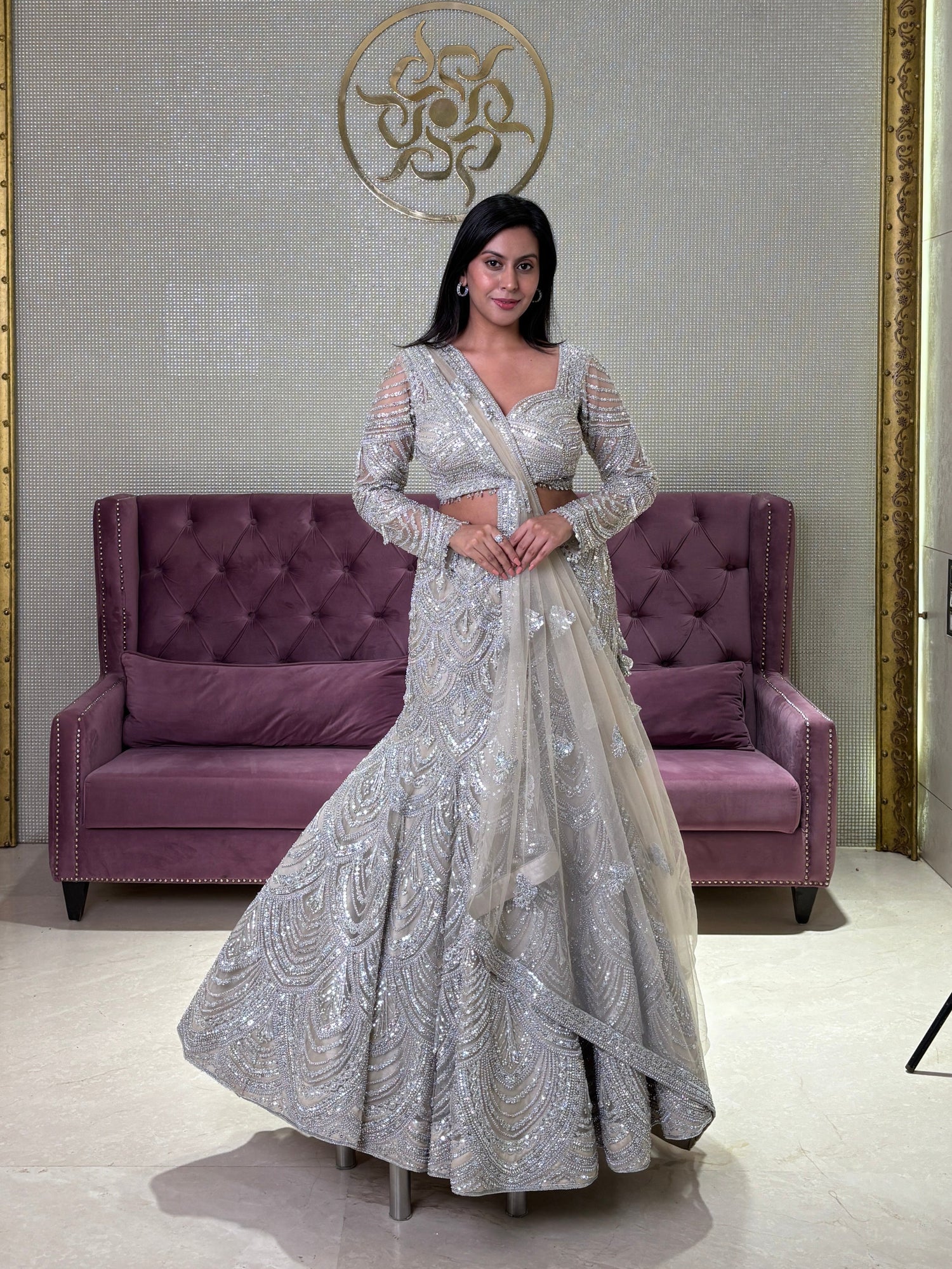 Woman in a grey net lehenga standing in a room with a purple couch and decorative wall.
