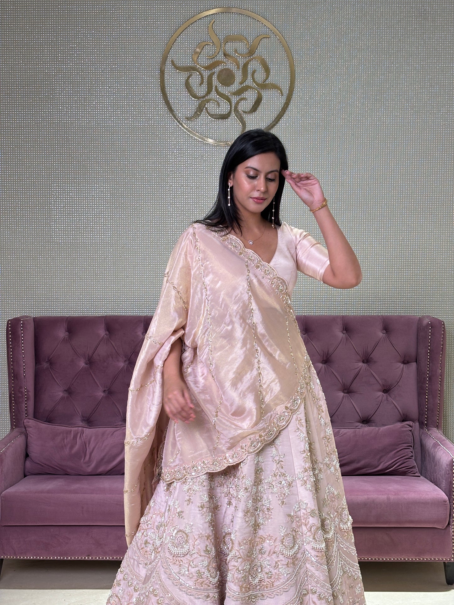 Woman in a long, light-colored pink lehenga standing next to a purple upholstered chair.