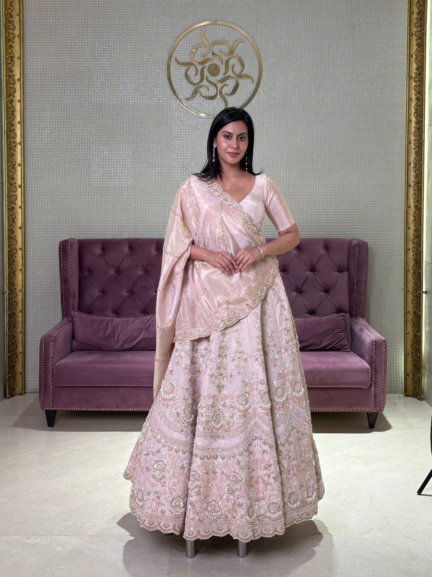 Woman in a pink tissue lehenga standing in a room with a purple couch and decorative wall.