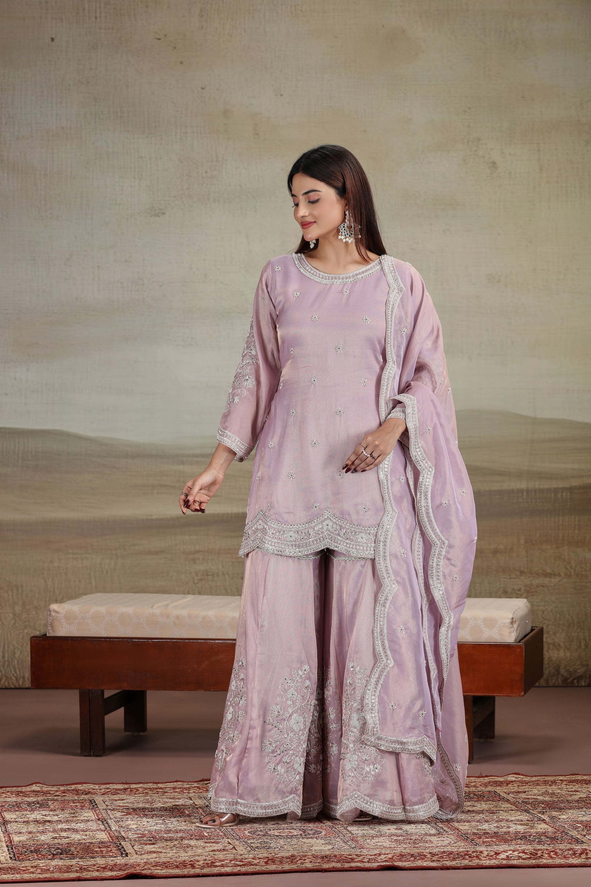 Woman in a lilac purple suit set standing in a room with a textured wall and wooden bench.