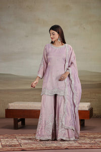 Woman in a lilac purple suit set standing in a room with a textured wall and wooden bench.